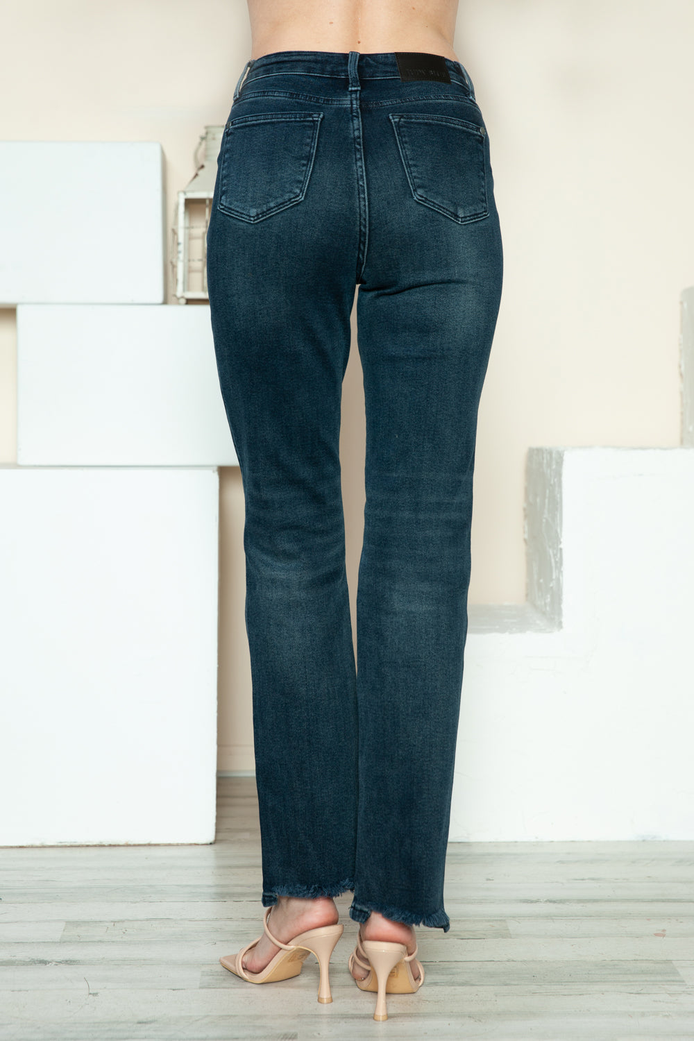 A person is modeling the Judy Blue Full Size Button Fly Hem Destroy Straight Jeans, high-waisted with distressed detailing and frayed hems, paired with light-colored open-toe heels. The background includes white blocks and a light-colored wall.