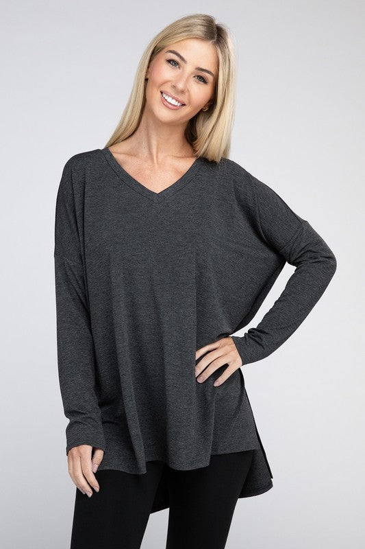 A woman with long, blonde hair is wearing a loose-fitting, brown Dolman Long Sleeve V-Neck Side Slit Hi-Low Hem Top and black pants. She poses with one hand on her hip and a neutral facial expression against a light grey background.