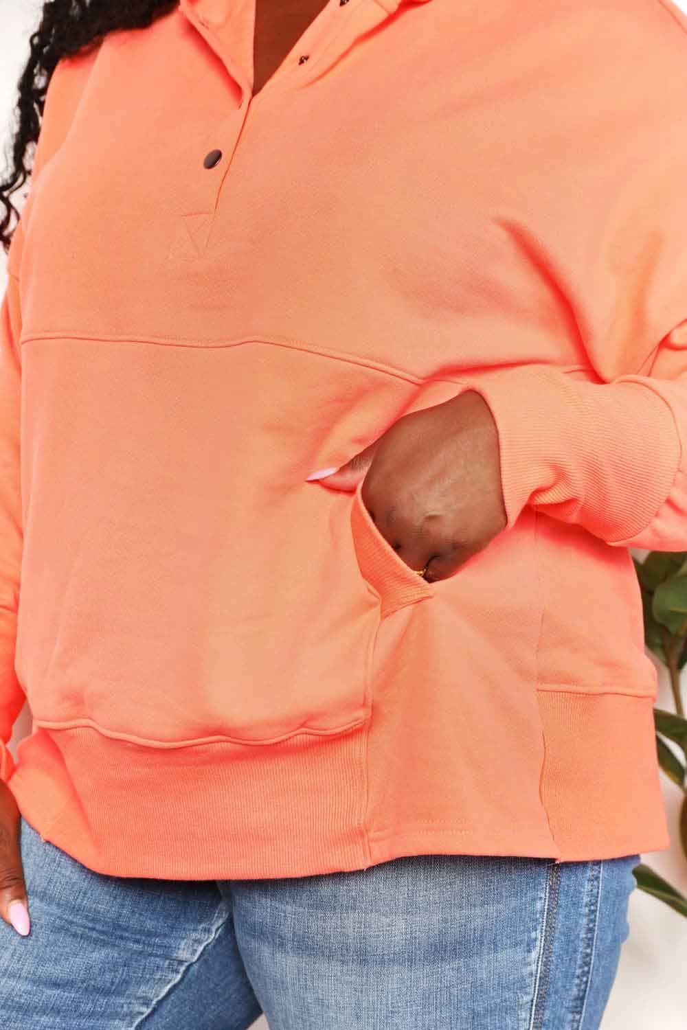 A woman with long hair wears a Double Take Quarter-Snap Dropped Shoulder Hoodie in solid orange. She is standing, with one hand touching her hair and the other resting in the pocket of her jeans, exuding a casual vibe.