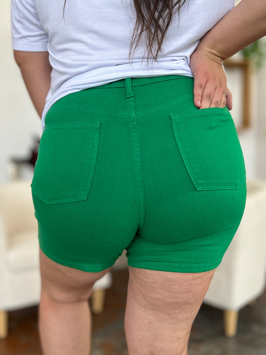 Two women stand indoors wearing white tops and Judy Blue Full Size Tummy Control Garment Dyed Denim Shorts in green. The woman on the left has her hand in her pocket, and both are smiling. There are plants and decorations in the background.