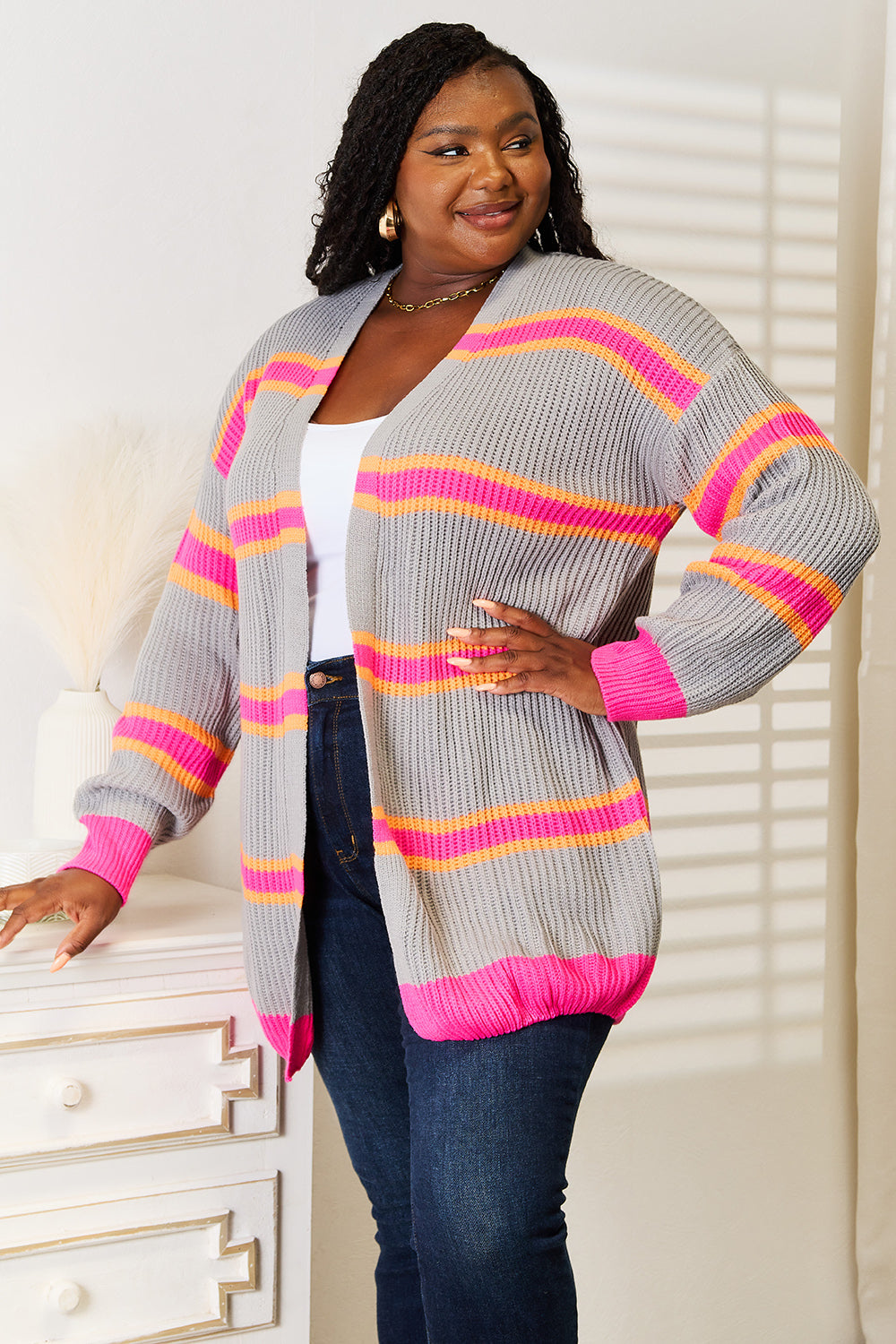 A woman stands against a white wall, smiling with one hand on her hip. She wears the Woven Right Ribbed Long Sleeve Cardigan, featuring pink and orange stripes, over a white top and dark jeans—an excellent choice for versatile layering.