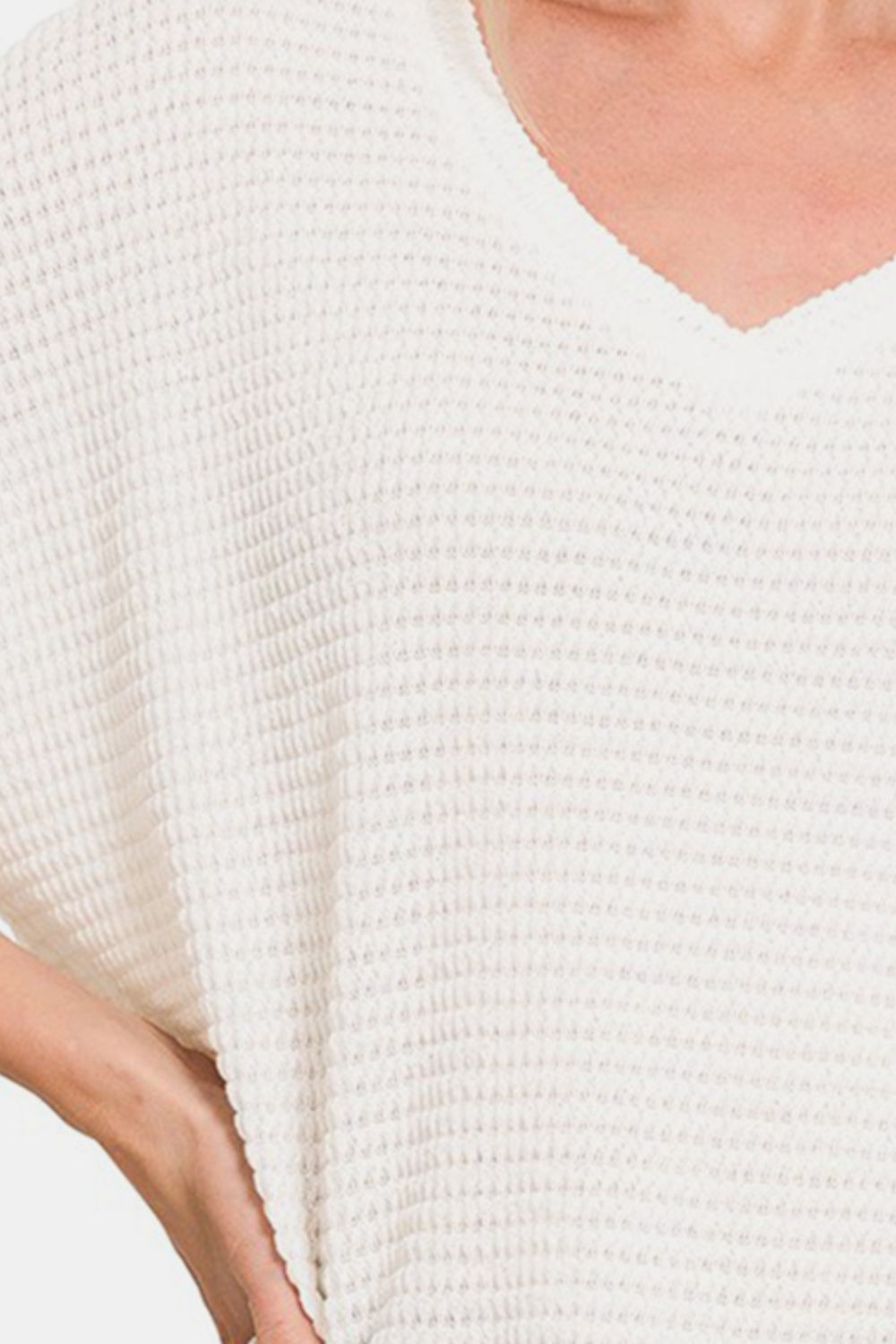 A woman wearing a Zenana Drop Shoulder Short Sleeve Jacquard Knit Top in white stands with one hand on her hip and smiles. She has long, light brown hair and is paired with dark blue jeans.