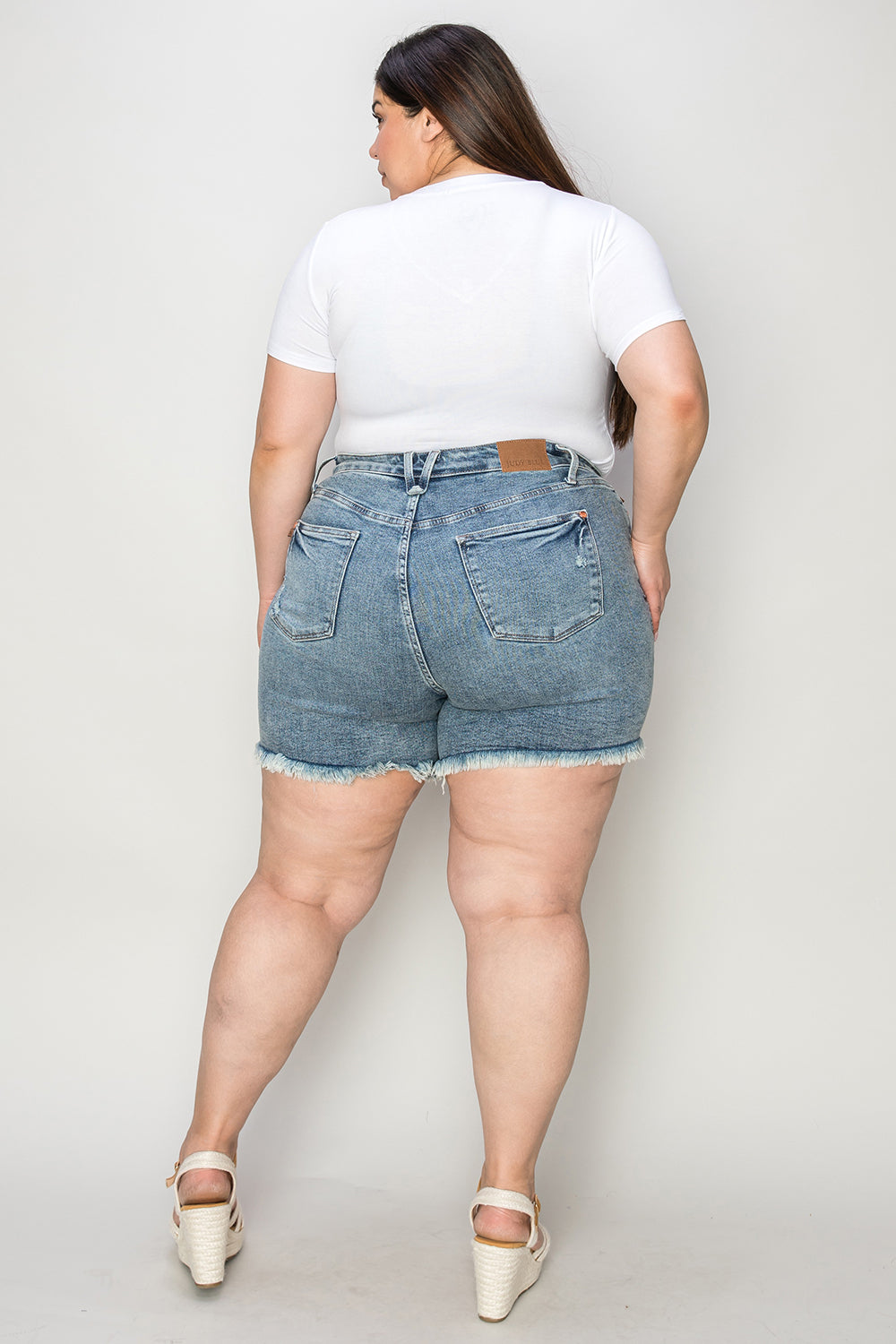 Wearing the Judy Blue Full Size Button Fly Raw Hem Denim Shorts and a white t-shirt, a person stands smiling in sandals against a plain background.