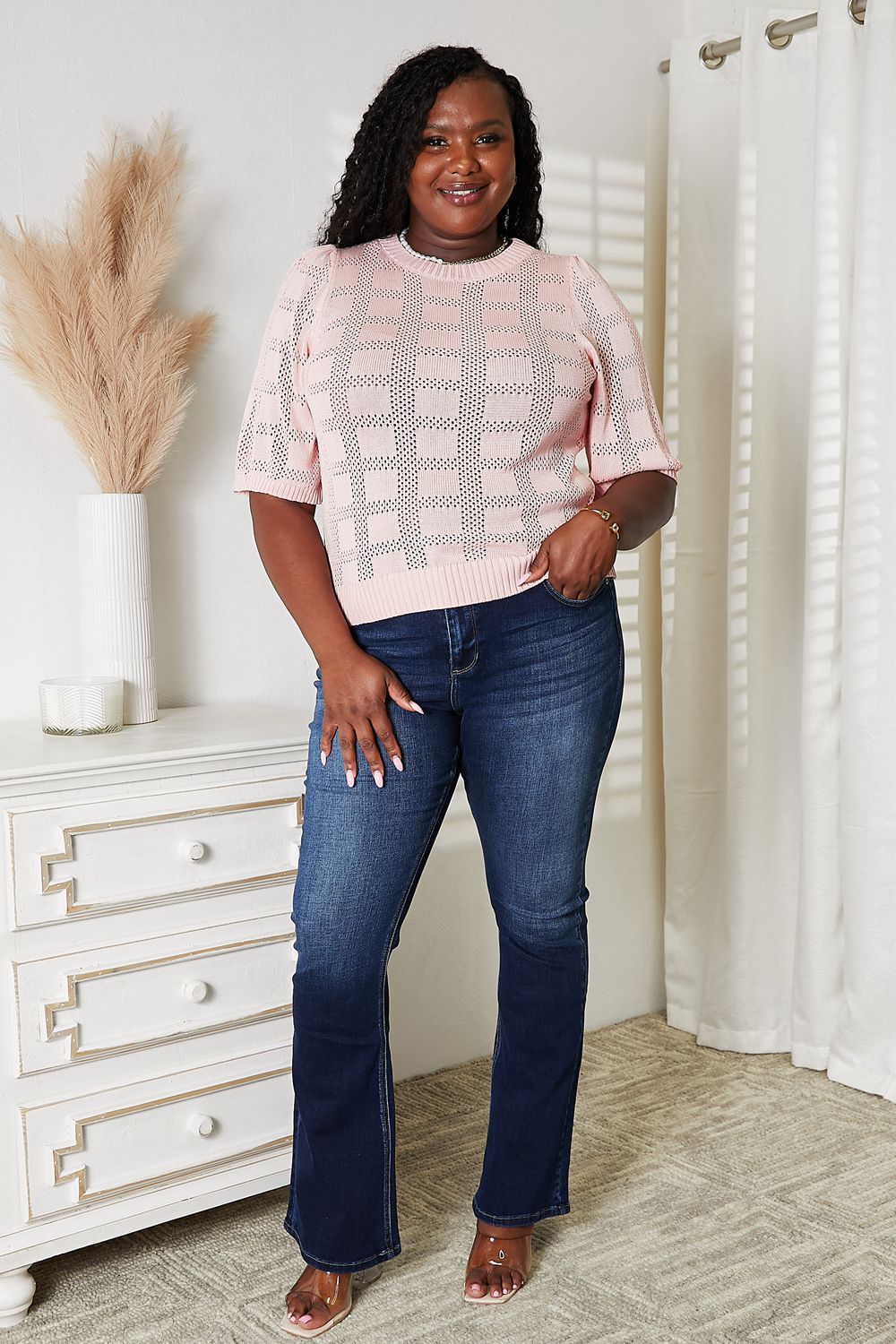 A person is standing and smiling, wearing the Mandy Ribbed Trim Round Neck Knit Top in light blue with half sleeves, paired with blue jeans. A potted plant is partly visible next to them against the plain background.