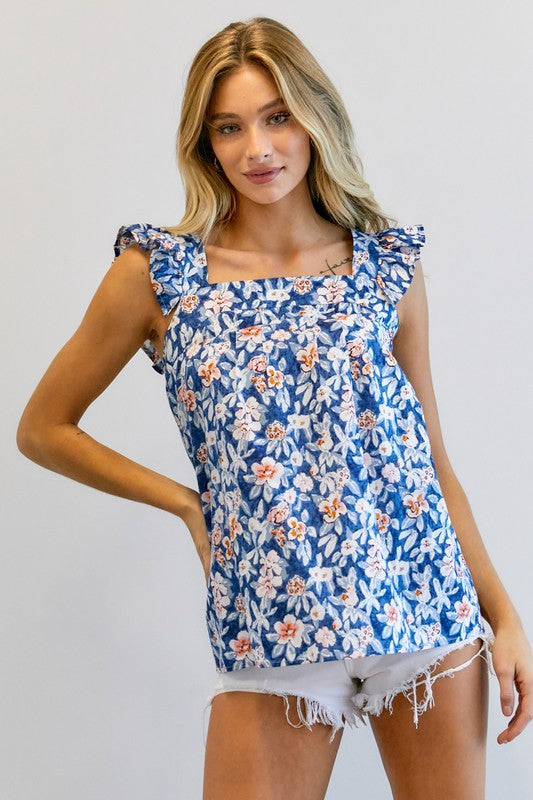 A woman in a vibrant summer wardrobe stands against a plain background, wearing the Floral Printed Ruffle Sleeveless Top paired with white shorts, with one hand casually tucked in her pocket.