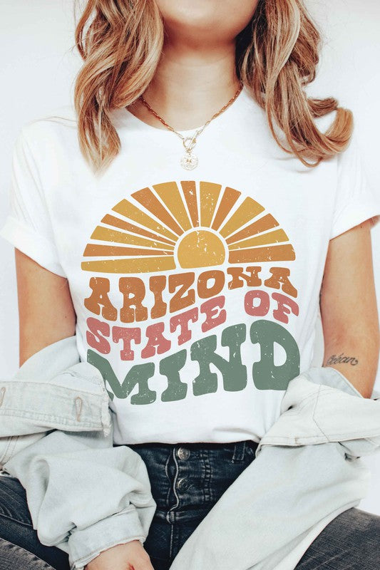 Person wearing the ARIZONA STATE OF MIND Graphic Tee, a beige 100% cotton t-shirt with "ARIZONA STATE OF MIND" text in brown and green colors. The unisex shirt offers both comfort and style for any occasion. They are holding a pink polka-dotted mug.