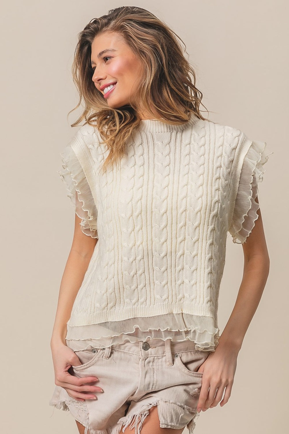 A woman is modeling the BiBi Layered Pleat Chiffon Edge Cable Knit Top, which features a cream-colored, sleeveless design. She wears it over a sheer top with layered pleat chiffon edges as she poses against a neutral background, looking to the side and smiling.