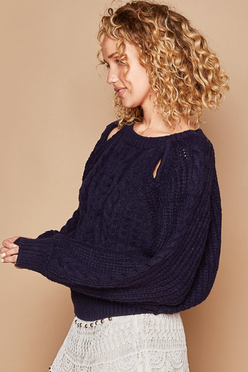 A person is wearing the POL Cable Knit Cutout Long Sleeve Sweater in navy, paired with a white crochet skirt, and is standing against a beige background.