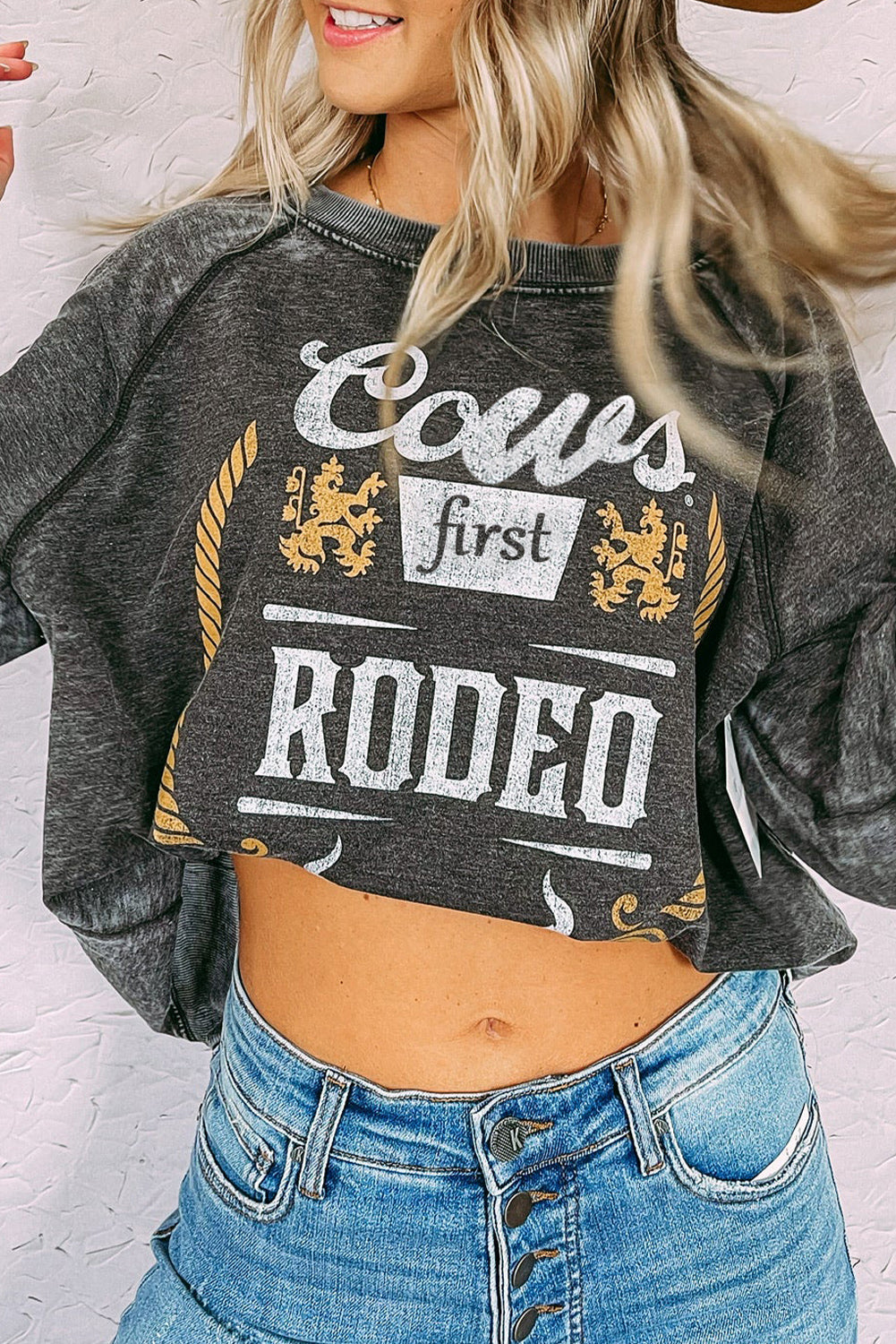 A blonde woman wearing a Gray Coors Banquet RODEO Graphic Mineral Washed Sweatshirt and light blue denim shorts, showcasing casual outdoor wear, stands outdoors with her back to the camera.