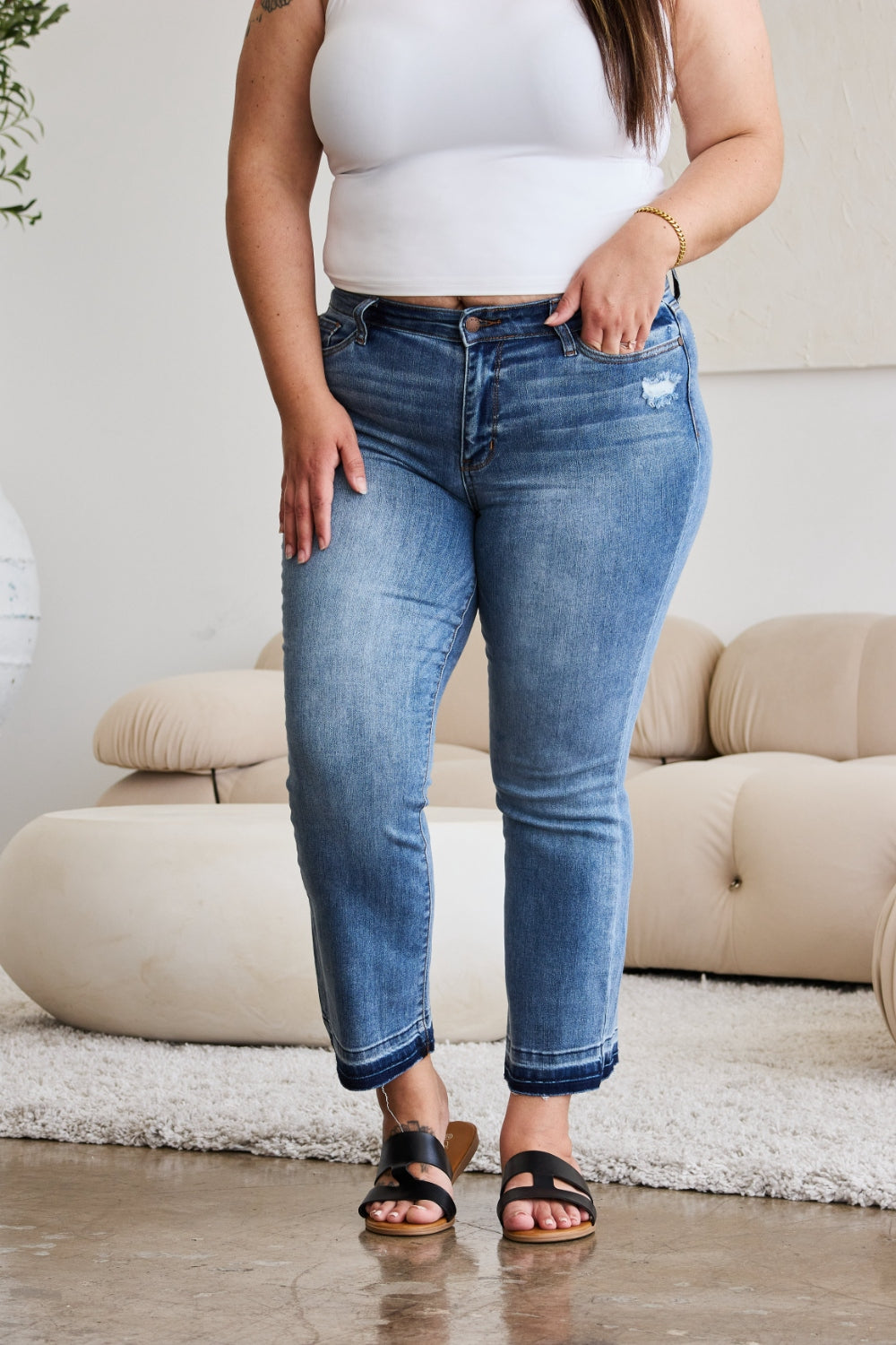 A person modeling Judy Blue Full Size Release Hem Cropped Bootcut Jeans and white heeled sandals stands in a room with light-colored furniture.