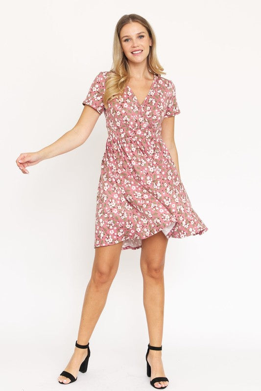 A woman wearing a short-sleeved, V-neck, Ditsy Surplice Fit and Flare Midi Dress with a dark floral background, crafted from luxurious Polyester Spandex and proudly Made in the United States, poses against a white backdrop, smiling as she softly holds the sides of her dress.