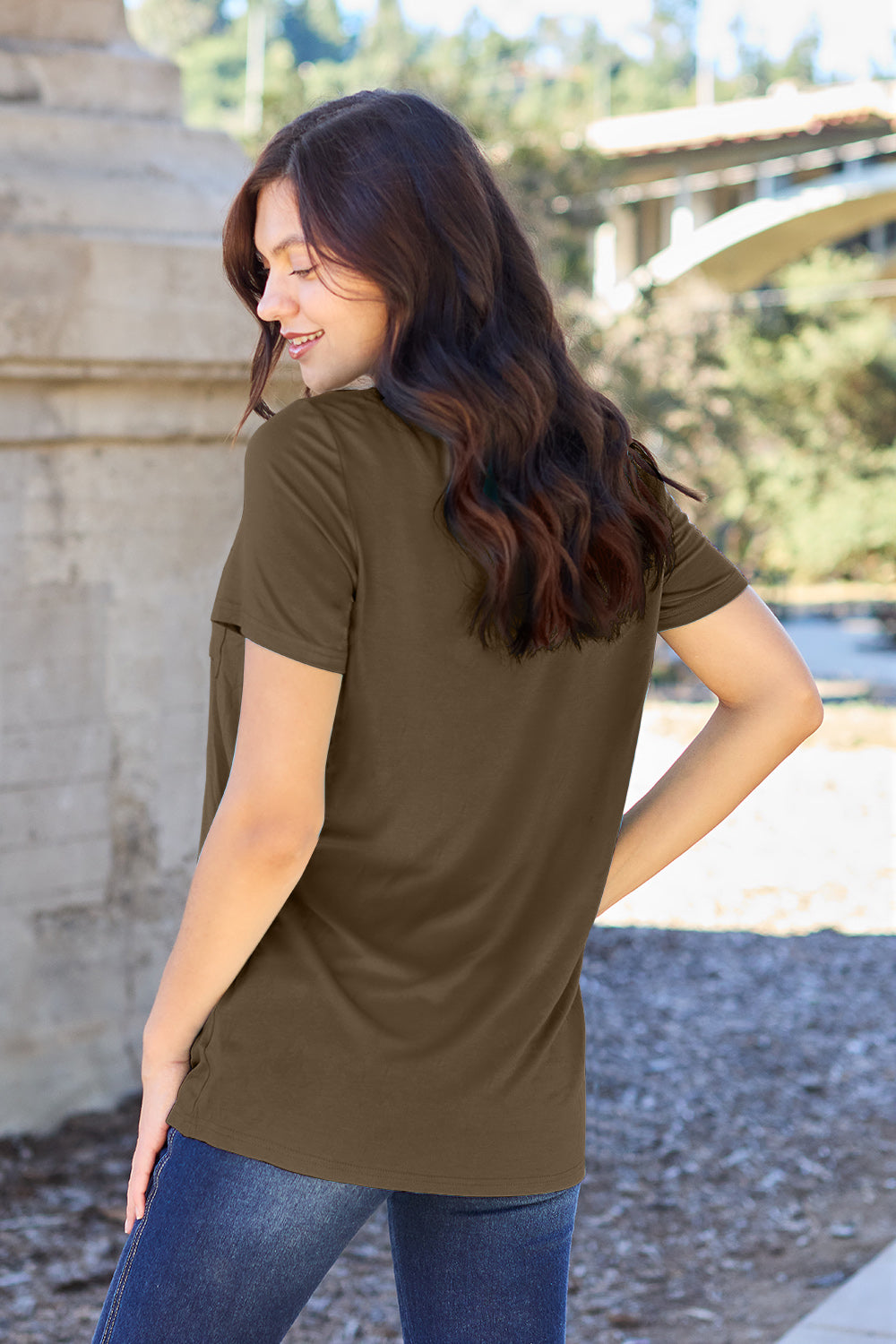 A woman with long brown hair, wearing a Basic Bae Full Size V-Neck Short Sleeve T-Shirt in teal and stretchy blue jeans, stands outdoors near a stone structure, smiling at the camera with one hand in her pocket.