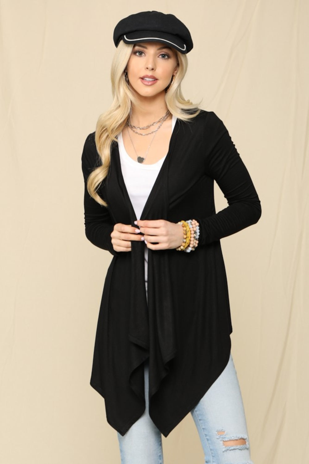 A woman wearing a black hat, the Celeste Full Size Open Front Knit Cardigan—an essential wardrobe staple—and a white top, holding a beige clutch, poses against a beige background.