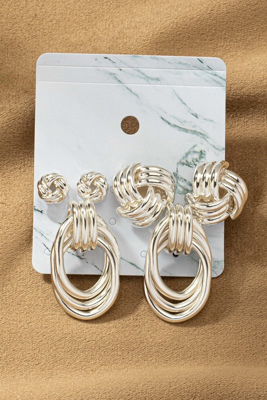 The Premium Trio Metal Knot and Hoop Earrings, displayed on a marble-patterned card, showcase three pairs of unique woven knot designs all embellished with shiny gold plating against a tan fabric backdrop.