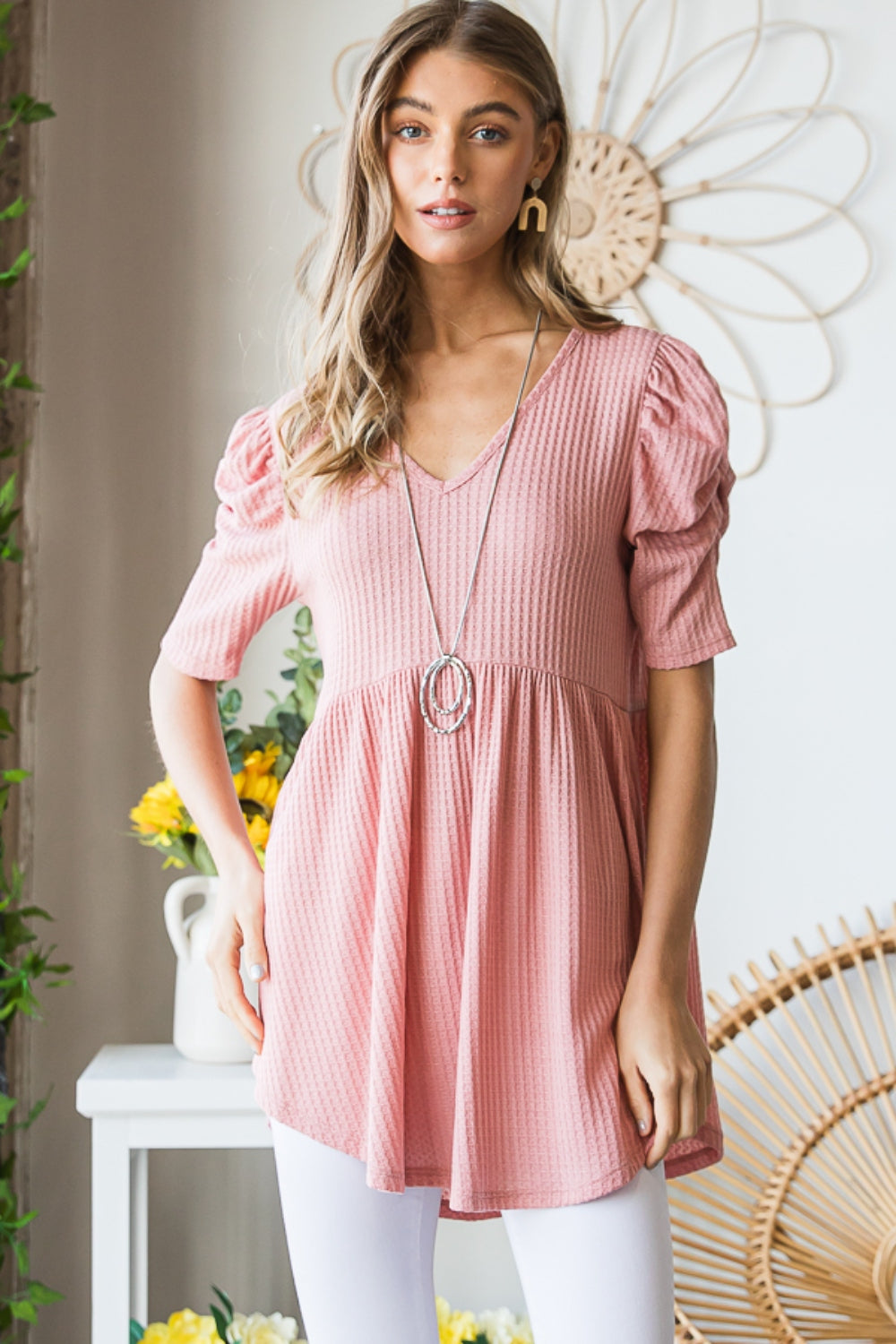 A woman in a Heimish Full Size Waffle Knit V-Neck Babydoll Top with short-sleeves and a long necklace, standing by a window with a neutral expression; the perfect cozy wardrobe addition.