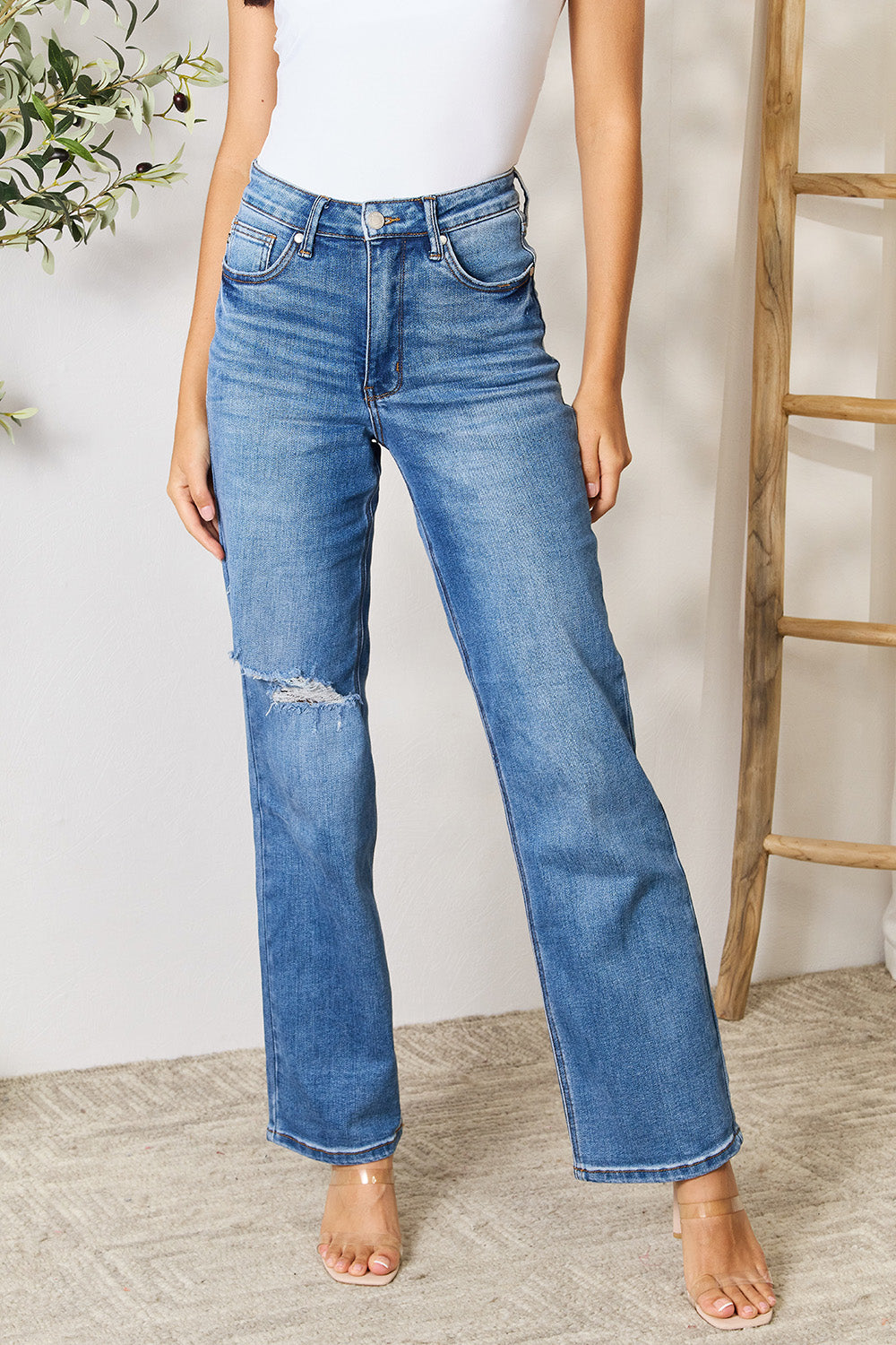 A person wearing a white top and Judy Blue Full Size High Waist Distressed Jeans stands in a room with a ladder and a plant.