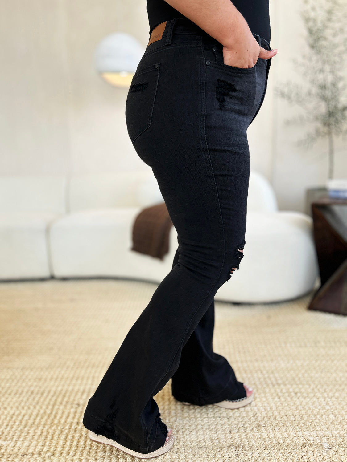 A person stands indoors, hands in pockets, wearing a white top and the Judy Blue Full Size High Waist Distressed Flare Jeans that create a flattering silhouette. In the background, a beige sofa and potted plants add to the retro-inspired look.