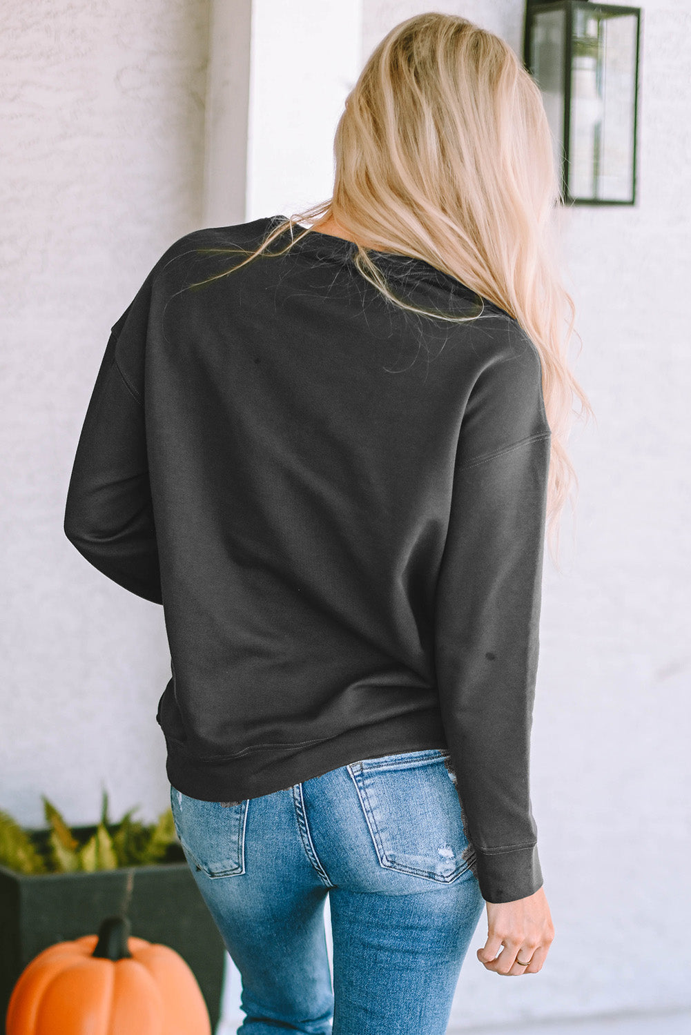 A person with long blond hair wearing a gray COOL MOMS CLUB drop shoulder sweatshirt and relaxed-fit blue jeans is seen from the back, standing near a pumpkin and a window.