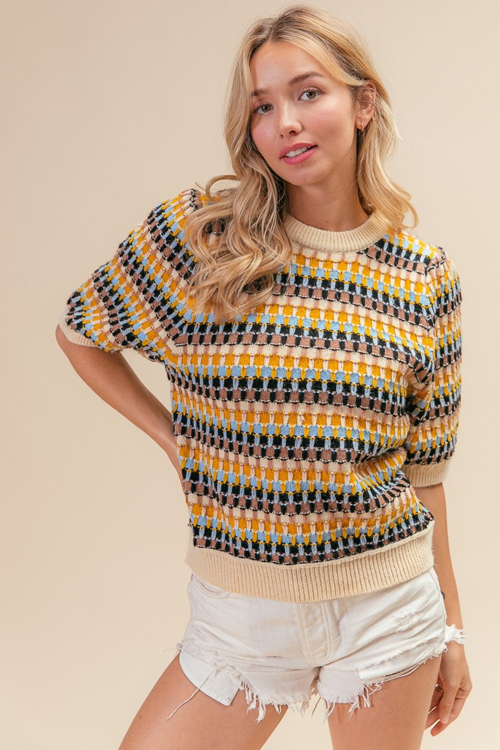 A woman with long blonde hair wearing a BiBi Multi Color Half Sleeve Sweater and white distressed shorts poses with one hand behind her head against a plain beige background.