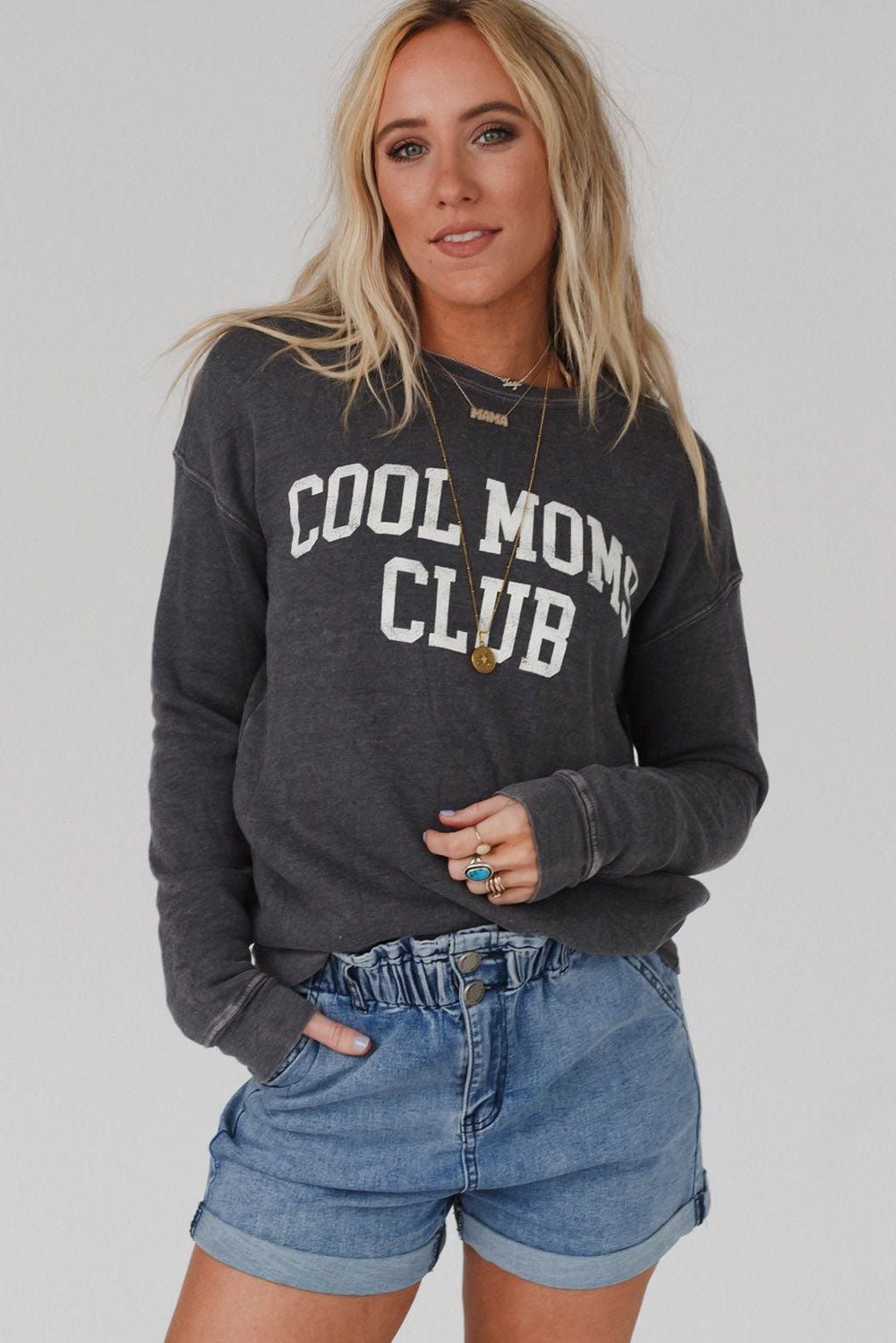 A person with long blond hair wearing a gray COOL MOMS CLUB drop shoulder sweatshirt and relaxed-fit blue jeans is seen from the back, standing near a pumpkin and a window.