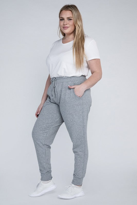 A person stands against a white background, wearing a white t-shirt, grey Plus-Size Jogger Pants with an adjustable waistband, and white sneakers. They have long blonde hair and are posing with one hand on their hip.