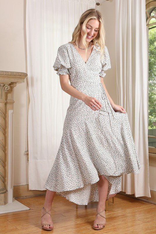 A woman with long blonde hair is wearing the SS V neck wrap dress in a green floral pattern, standing in front of a neutral background and smiling.