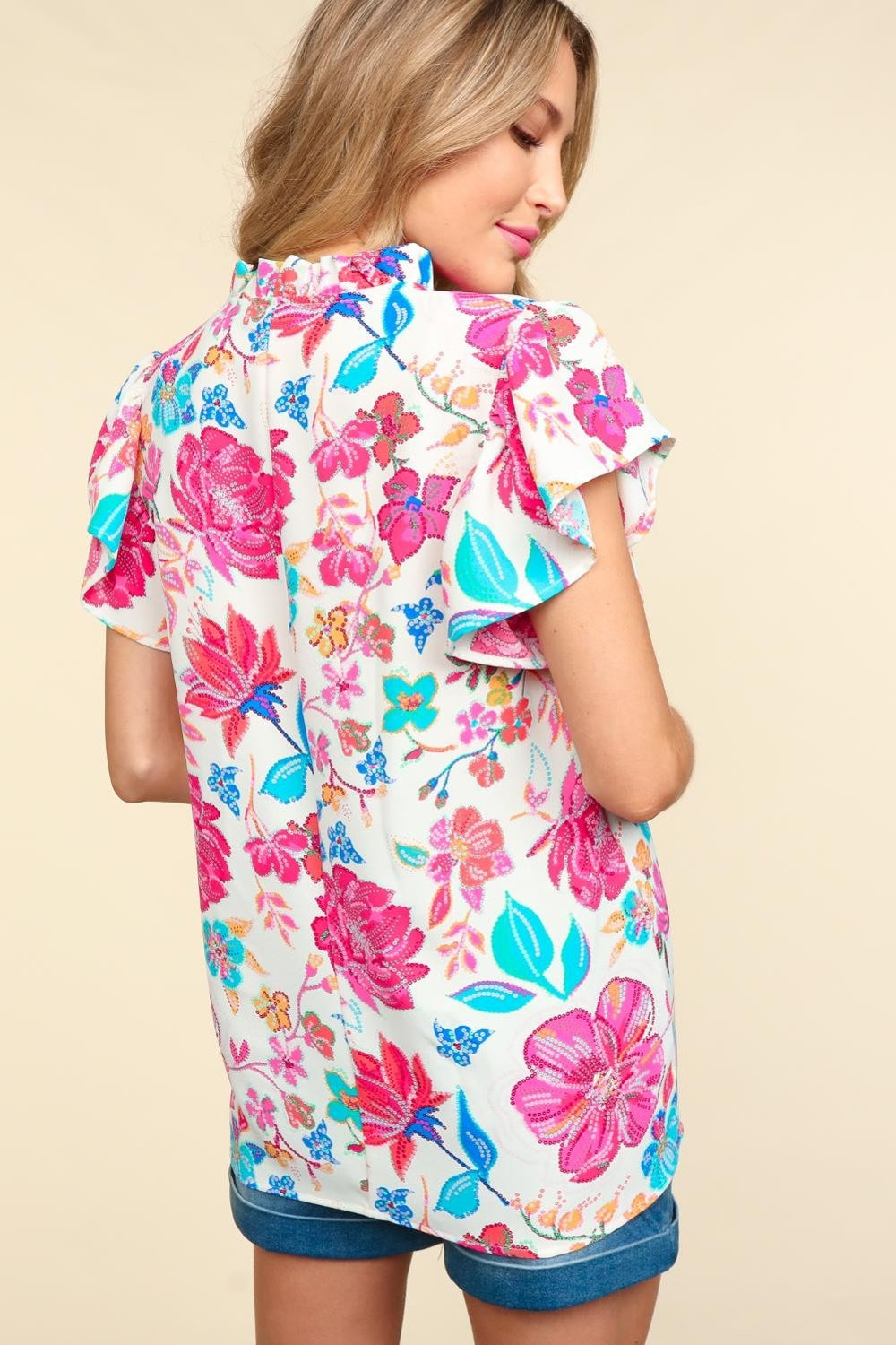 A woman wearing the Haptics Notch Neck Sequin Floral Blouse, adorned with elegant pink, red, and blue flowers, stands against a beige background.