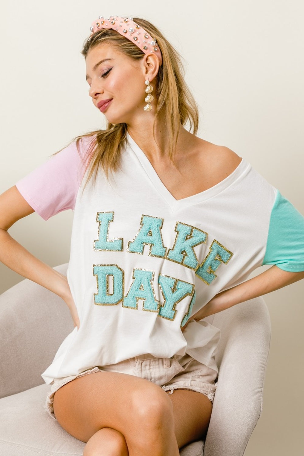 A woman wearing a BiBi Letter Patch V-Neck Color Block Short Sleeve T-Shirt, which features "Lake Days" printed on it, sits on a chair. She holds the shirt's sleeves and wears a headband and earrings.