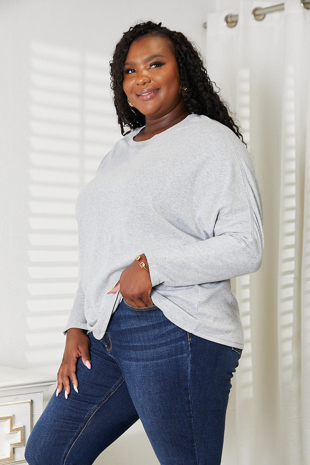 A person with long hair wearing a Double Take Seam Detail Round Neck Long Sleeve Top in solid black and blue jeans stands with one hand in their pocket, looking downward and smiling.