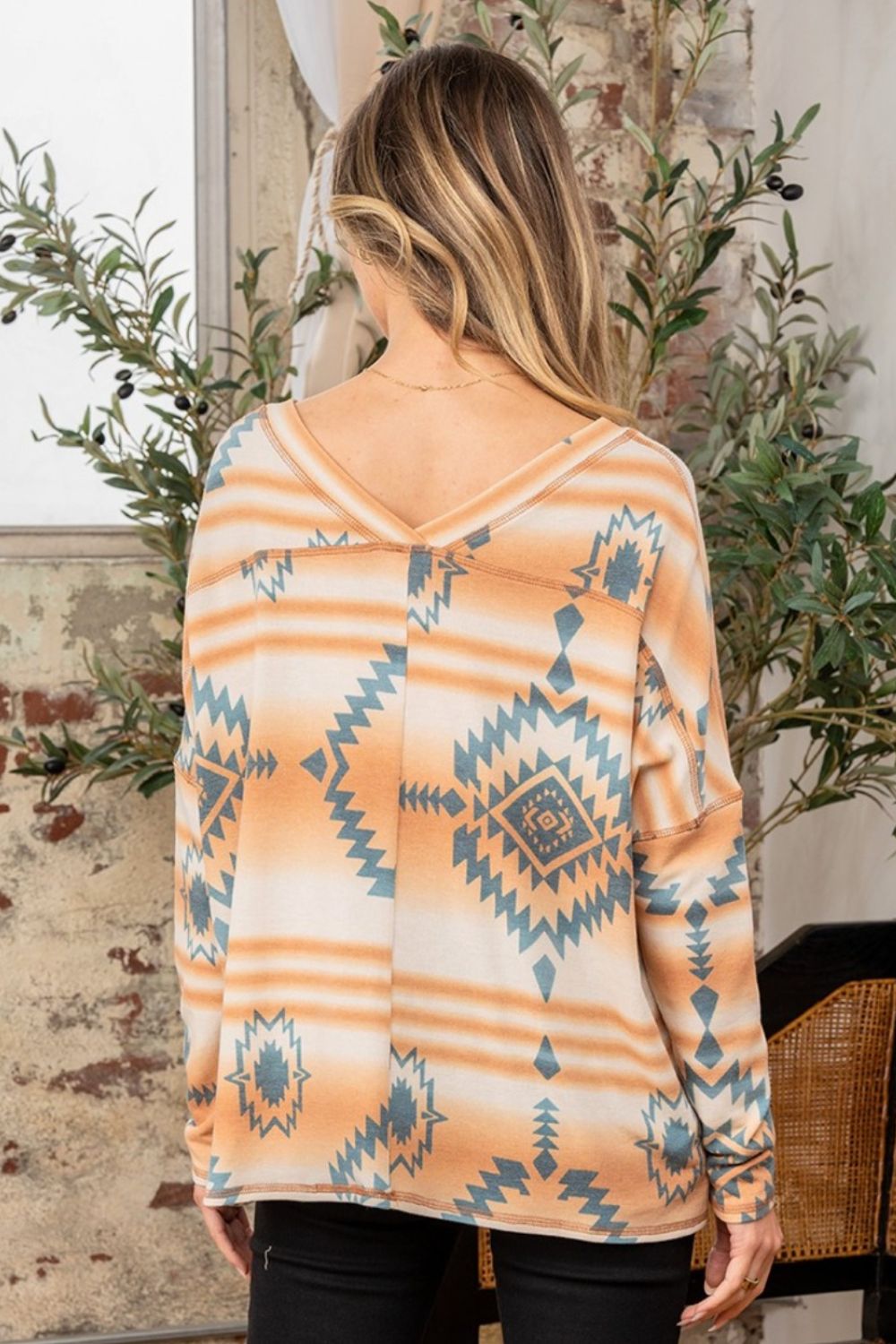 A woman stands indoors wearing the Sew In Love Full Size Printed French Terry V-Neck T-Shirt, adorned with geometric designs. She is posed with one hand resting on a chair, and plants are visible in the background.