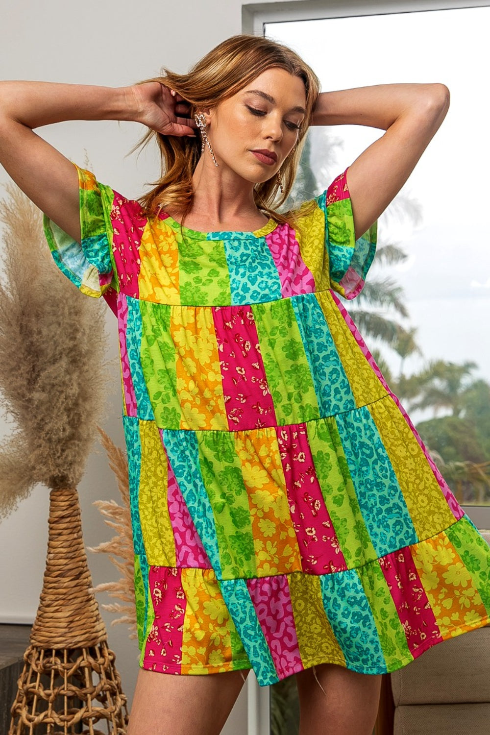 A woman in a BiBi Leopard Short Sleeve Tiered Dress poses with her arms raised behind her head. She stands indoors near a window, framed by pampas grass decor, exuding effortless style and grace.