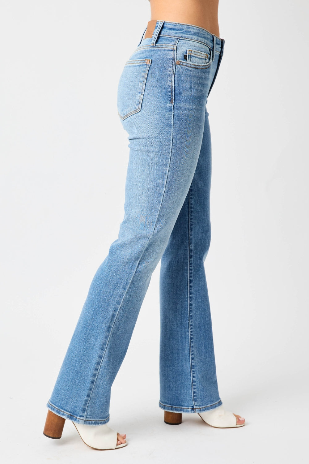 Person wearing Judy Blue Full Size Mid-Rise Waist Straight Jeans and a black top, standing in a room with white chairs, potted plants, and wooden shelves. Only the lower half of the body and part of the arms are visible.