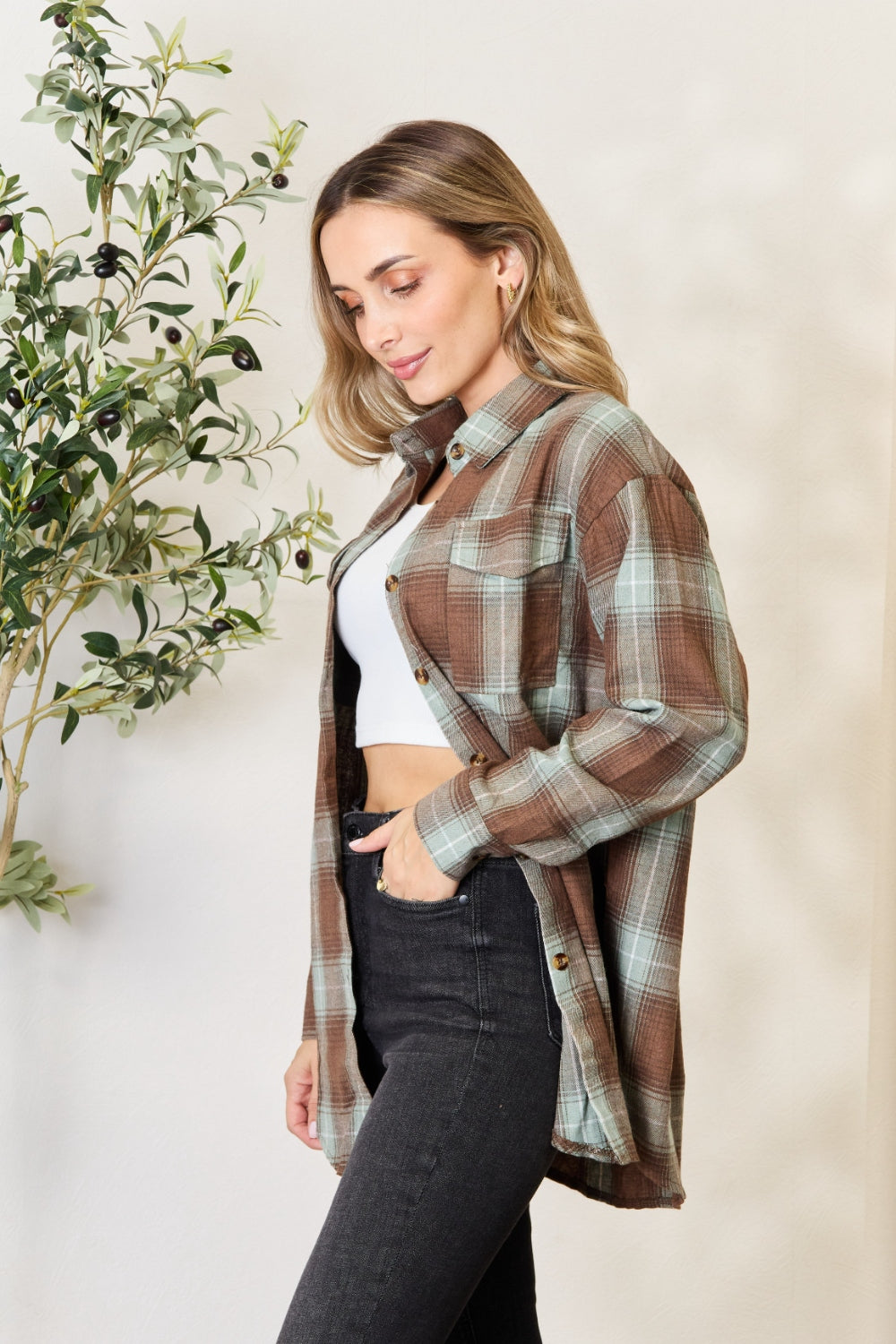 A woman stands in front of a light-colored wall and an olive tree, wearing the Mandy Plaid Dropped Shoulder Shirt paired with a white crop top and dark jeans. She smiles and holds her hair back with one hand.