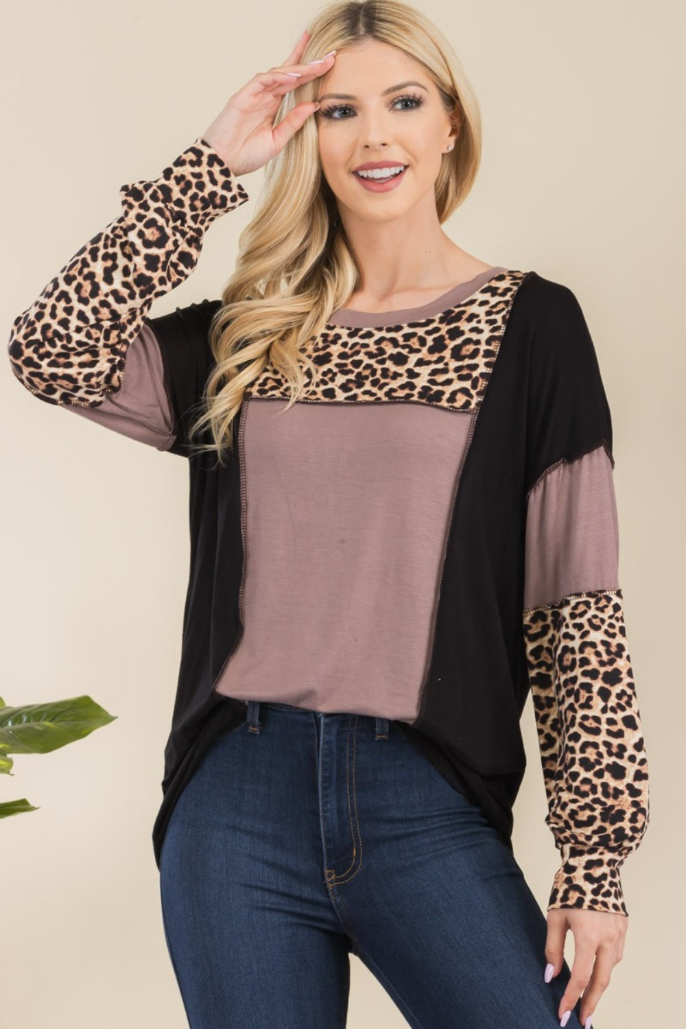 A person wearing the Celeste Full Size Leopard Color Block Exposed Seam T-Shirt and dark jeans stands against a neutral background with greenery. The long-sleeved shirt, with its stylish exposed seams, features cozy leopard print accents, adding subtle flair to this chic ensemble.