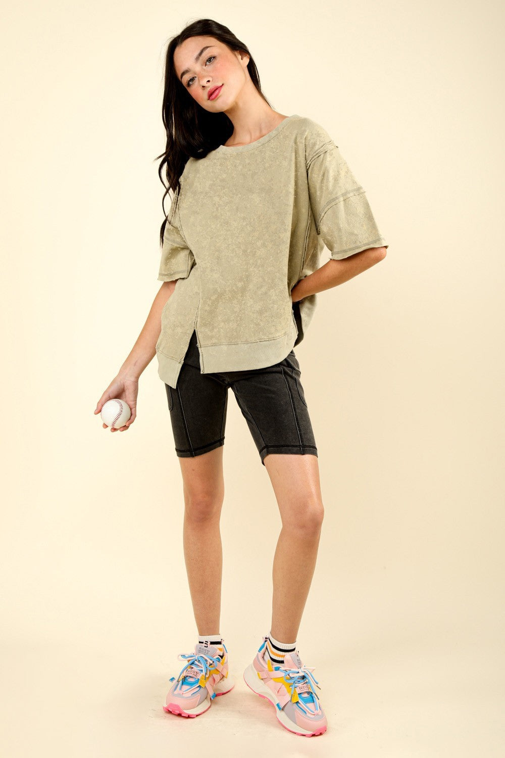 A woman with long dark hair stands holding a baseball. She is wearing the VERY J Round Neck Exposed Seam Slit T-Shirt and black shorts. The background is plain beige.
