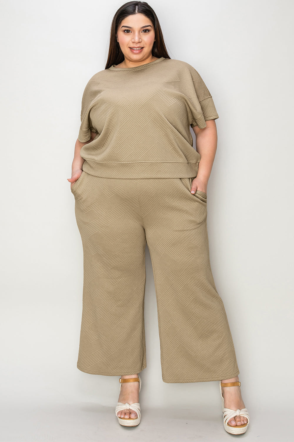 A woman with long hair smiles while standing outside a building, dressed in the Double Take Full Size Texture Short Sleeve Top and Pants Set, featuring a matching beige two-piece ensemble. She wears slides and holds a hat in her right hand. Her outfit, slightly stretchy for added comfort, is perfect for machine wash cold.