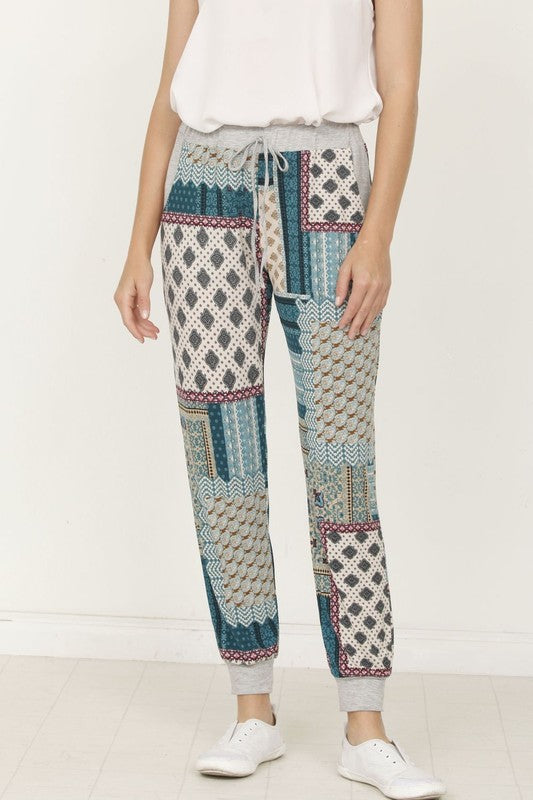 A person is standing indoors, with only part of their upper body visible, wearing a white top and Plus Quilted Print Joggers featuring a colorful pattern. The joggers have a drawstring waist and pockets where the person's hands are tucked. Their left foot is bare.
