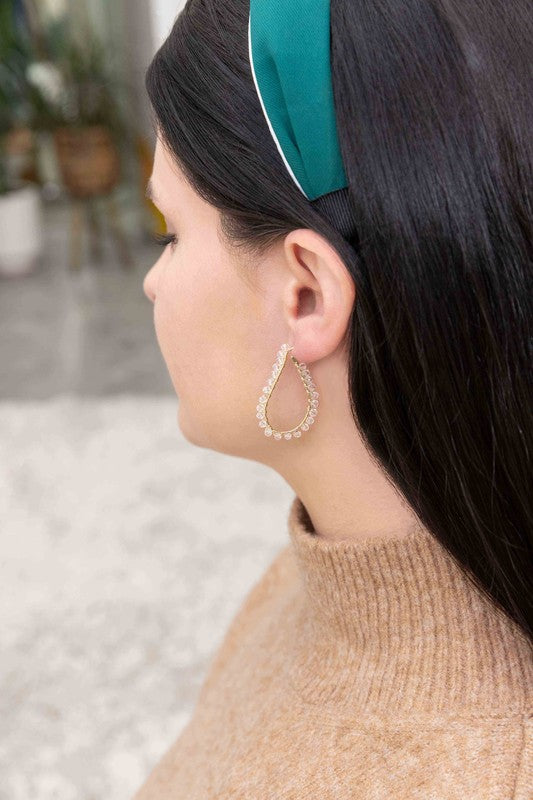 The Delicate Drops Hoop Earrings, featuring gold teardrop designs with pearl accents and clear beads, are displayed on a white surface.