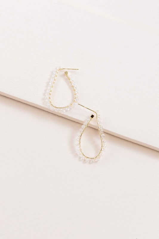 The Delicate Drops Hoop Earrings, featuring gold teardrop designs with pearl accents and clear beads, are displayed on a white surface.