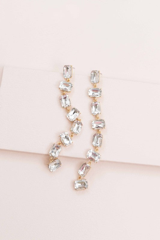 More Drama Dangle Earrings with clear rectangular diamond stones on a pastel pink background, perfect for formal occasions.