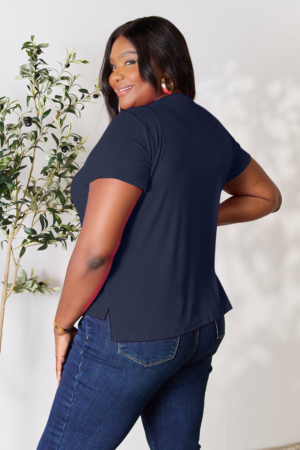 A person with wavy hair, wearing the Basic Bae Full Size Round Neck Short Sleeve T-Shirt in bright pink and beige pants, stands near a plant, smiling and looking to the side. The outfit embodies a basic style and offers slightly stretchy comfort, ideal for those who value effortless elegance.