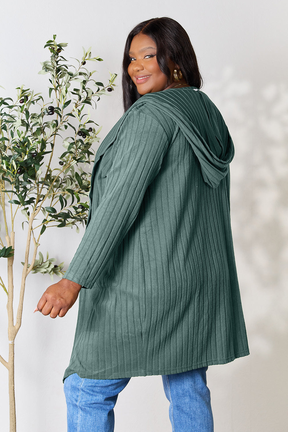 A woman wearing the Basic Bae Full Size Hooded Sweater Cardigan in black, paired with a white top and khaki pants, stands in front of a light background accented by a leafy plant on her left. With one hand tucked into her pocket, she gazes to her right.