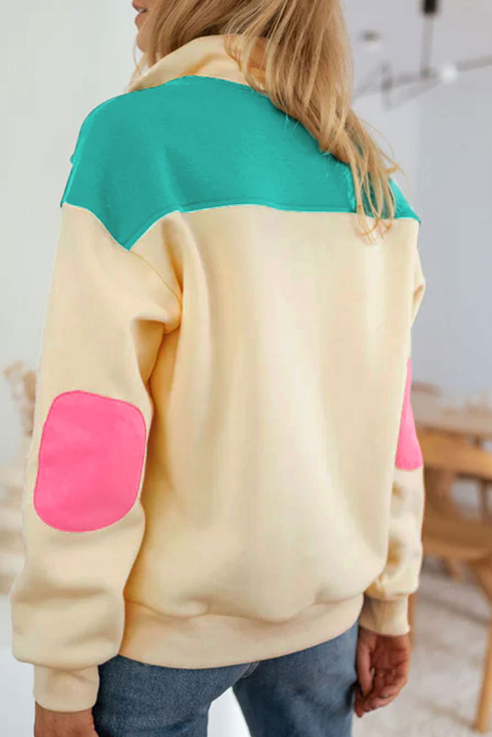 A woman wearing the Apricot Color Block Elbow Patch Half Button Sweatshirt, featuring cream and mint colors with pink elbow patches and a turn-down collar with snap button closure, stands outdoors. She has long wavy hair, sunglasses, a hat, and is wearing blue jeans.