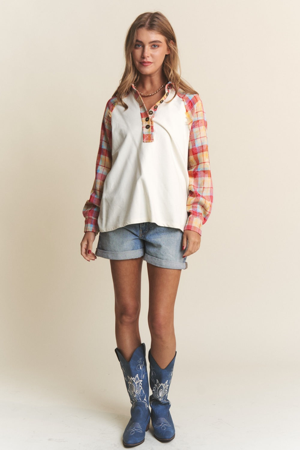 A woman wearing the J.NNA Plaid Contrast Button Down Henley Top—a stylish white pullover shirt with red and yellow plaid sleeves—and denim shorts, stands against a plain background.