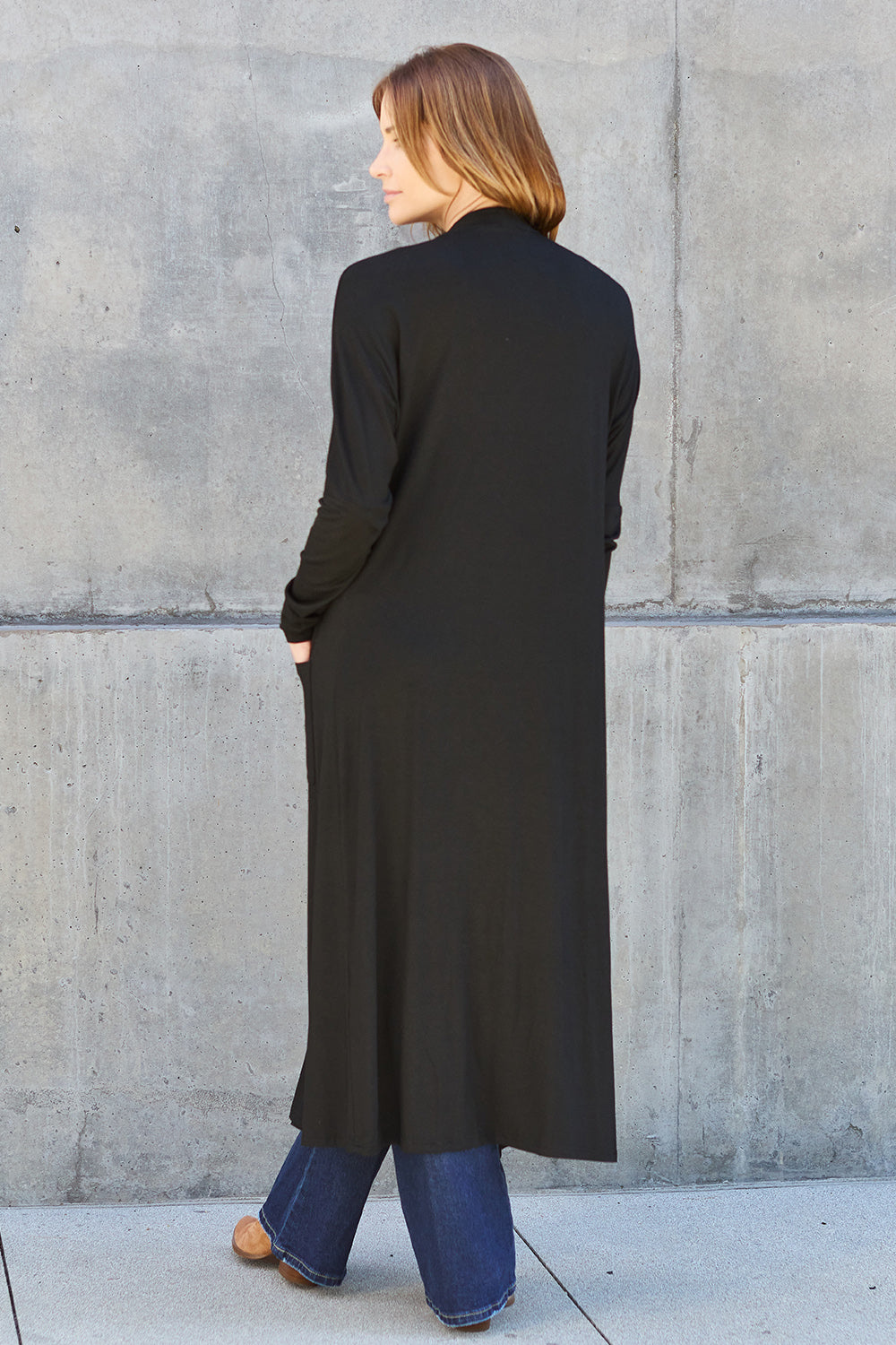 A woman stands against a concrete wall, wearing the Basic Bae Full Size Open Front Long Sleeve Cover Up made from rayon spandex, a white top, blue jeans in her perfect size, and brown shoes with her hands in her pockets. Machine wash cold for best results.