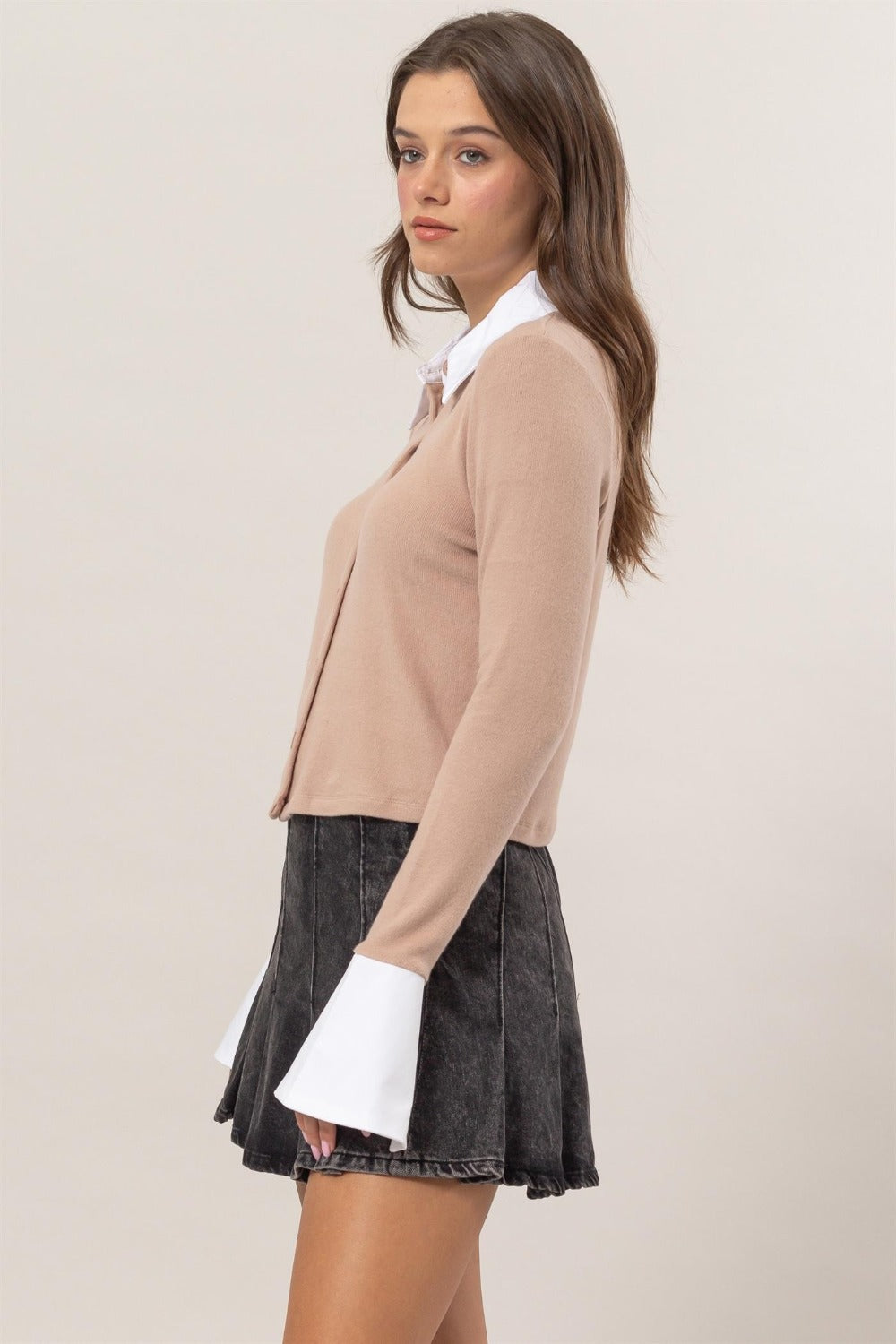 A woman stands wearing the HYFVE Collared Contrast Button Down Crop Knit Cardigan with white cuffs and collar, a dark pleated skirt, and black ankle boots. She poses with her hand on her hip in front of a plain background.