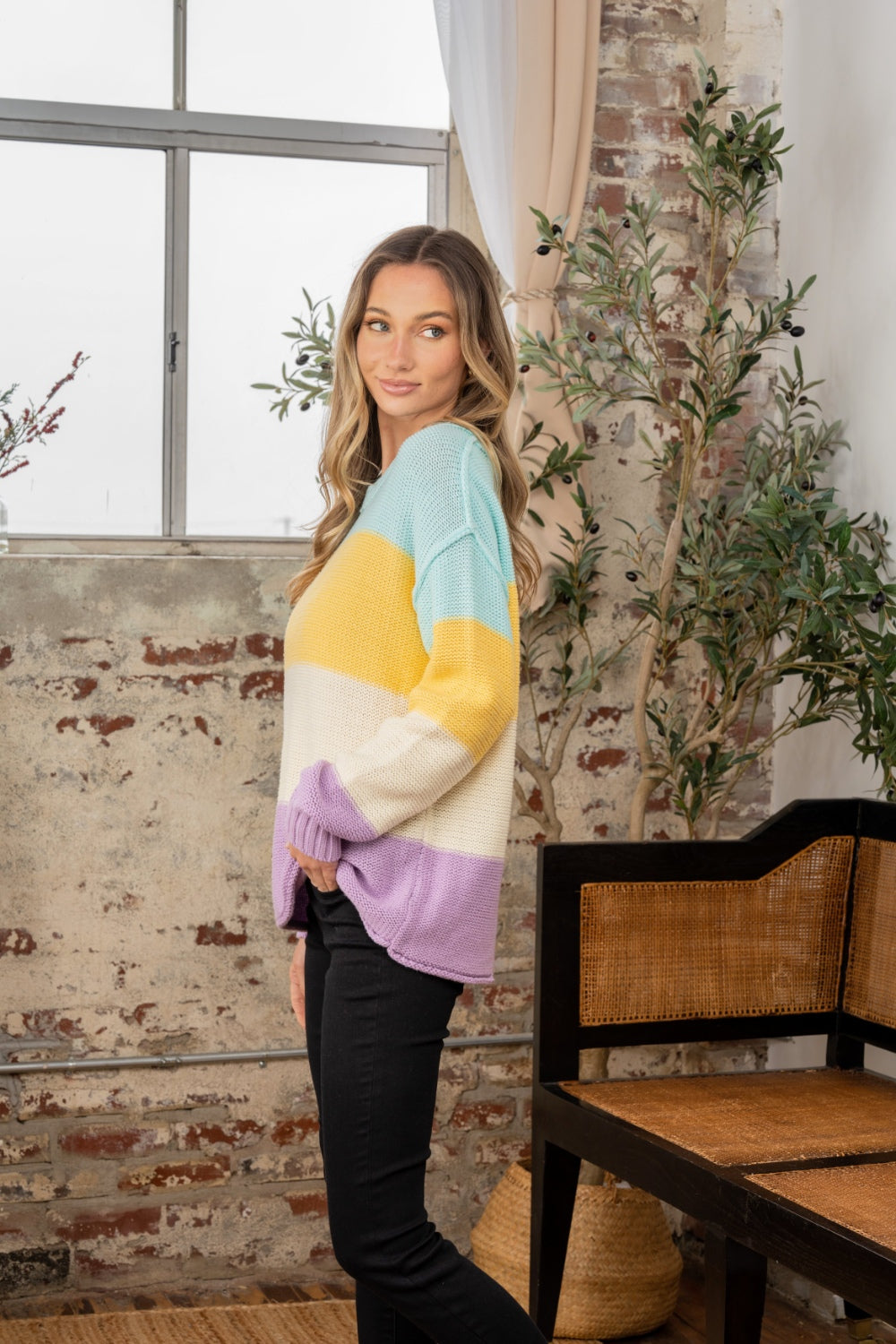 A person stands indoors near a window, wearing the Sew In Love Full Size Color Block Exposed Seam Sweater and black pants. The cozy knitwear complements the rustic walls and nearby plant, adding a touch of warmth to the scene.