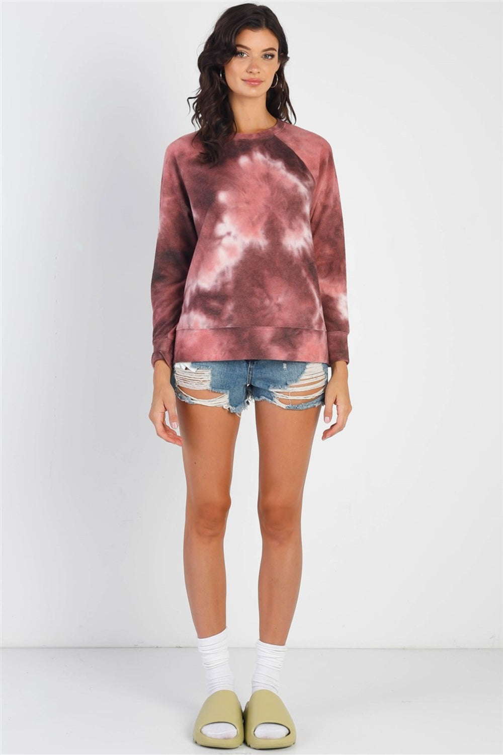 A person wearing the Cherish Apparel Tie-Dye Round Neck Long Sleeve Sweatshirt and distressed denim shorts stands against a plain background, showcasing effortless casual fashion.