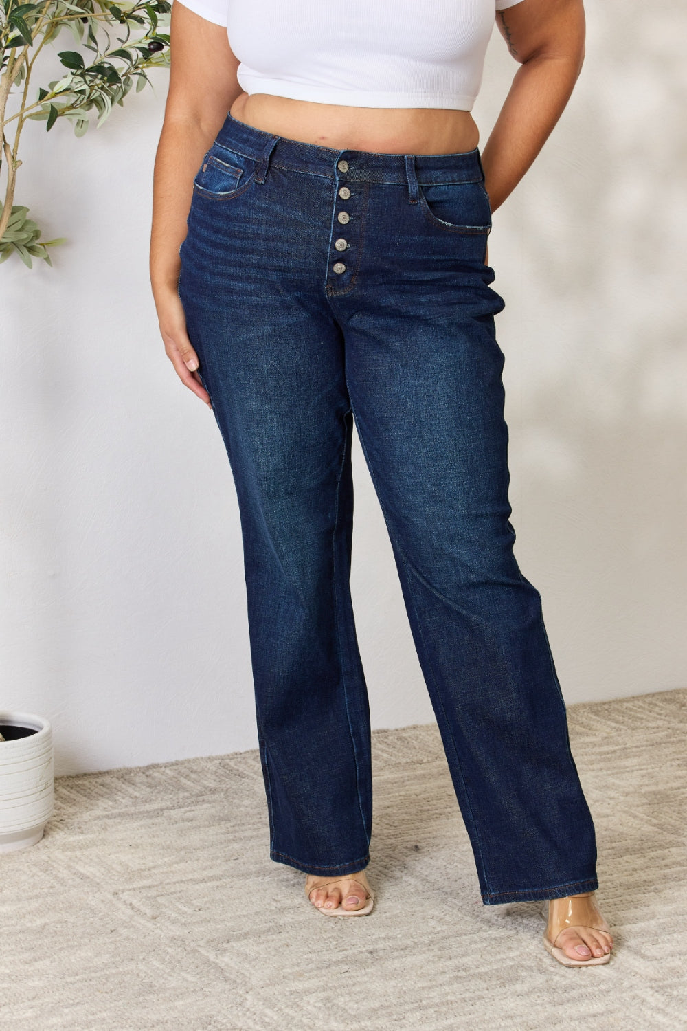 A person stands with hands in pockets, wearing the Judy Blue Full Size Button-Fly Straight Jeans, known for their high-waisted dark blue design crafted from premium lycra fabric. The jeans showcase button details and a straight leg cut, complemented by a white top and open-toed heels.