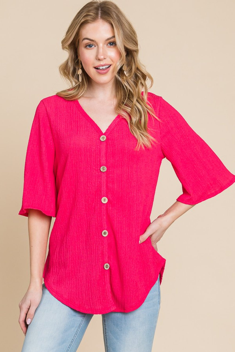 A person is wearing the BOMBOM Texture Decorative Button V-Neck Top, a chic design in bright pink featuring elegant details like short bell sleeves, and pairing it with blue jeans, stands against a beige background.